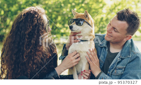 Cheerful young couple is having fun with dog putting sunglasses on its eyes and laughing while spending weekend in the park. Nature, modern lifestyle and animals concept. 95370327