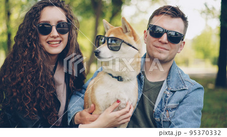 Portrait of cute couple and shiba inu dog all wearing sunglasses looking at camera and smiling sitting on grass in park. Modern lifestyle, cute animals and summer nature concept. 95370332