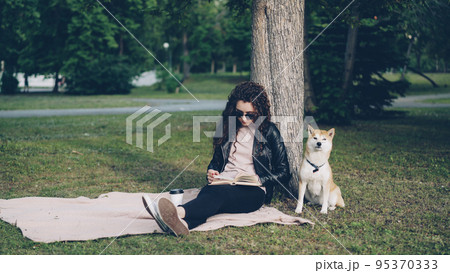 Relaxed young girl is reading book and drinking takeaway coffee sitting on lawn in park while her cute dog is smelling ground and air sitting under tree. Leisure and pets concept. 95370333
