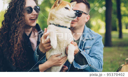 Joyful young people are playing with beautiful dog in the park having fun and laughing. Pretty girl with long curly hair is wearing leather jacket, guy is in modern denim jacket. Joyful young people are playing with beautiful dog in the park having fun and laughing. Pretty girl with long curly hair is wearing leather jacket, guy is in modern denim jacket. 95370402