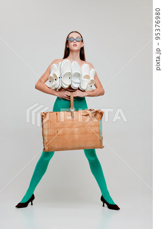 Portrait of young beautiful girl posing in green tights, paper bag and top made from slippers isolated on grey background. Unusual style Portrait of young beautiful girl posing in green tights, paper bag and top made from slippers isolated on grey background. Unusual style 95376980