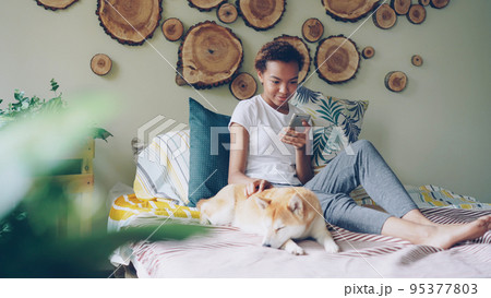 Pretty young woman is using smartphone and caressing lovely puppy lying on bed near its owner in modern style bedroom at home. Technology, domestic animals and people concept. Pretty young woman is using smartphone and caressing lovely puppy lying on bed near its owner in modern style bedroom at home. Technology, domestic animals and people concept. 95377803