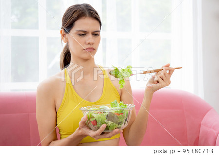 Young woman sitting on sofa eating vegetable salad while feeling dislike and expression disgusted. 95380713