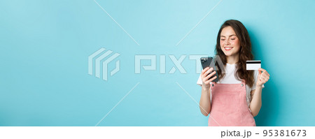 Young modern girl paying online with credit card, type info on smartphone, looking at screen with pleased face, shopping in app, standing over blue background Young modern girl paying online with credit card, type info on smartphone, looking at screen with pleased face, shopping in app, standing over blue background 95381673