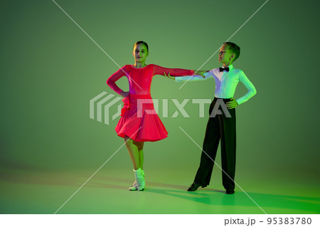 Beginner dancers. Portrait of beautiful little boy and girl dancing ballroom dance isolated over green background in neon light. Concept of art, beauty, grace, action, emotions. Beginner dancers. Portrait of beautiful little boy and girl dancing ballroom dance isolated over green background in neon light. Concept of art, beauty, grace, action, emotions. 95383780