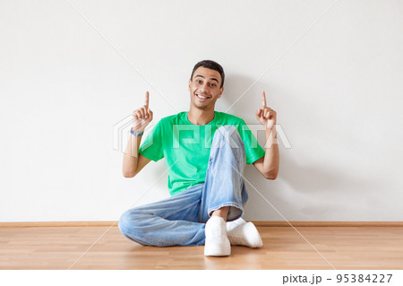 Joyful arab man sitting on floor and pointing at free space above him for ad, smiling at camera over white wall Joyful arab man sitting on floor and pointing at free space above him for ad, smiling at camera over white wall 95384227