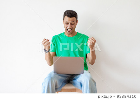 Promotion at work concept. Happy arab man celebrating success looking at laptop, sitting on chair over light wall Promotion at work concept. Happy arab man celebrating success looking at laptop, sitting on chair over light wall 95384310