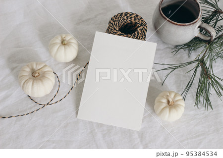 Christmas stationery. Closeup of empty greeting card, invitation mockup. Cup of coffee, pine tree branches and decorative rope. Linen tablecloth. White pumpkins Winter, Thanksgiving flatlay, top view. 95384534