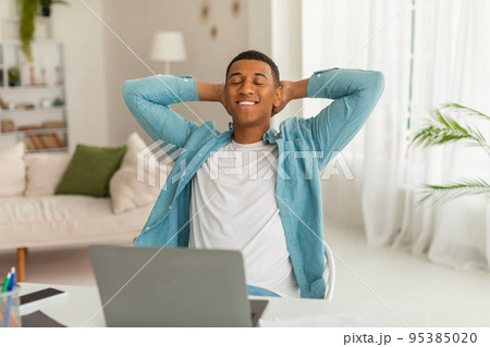 Cheerful millennial african american man with closed eyes resting at work with laptop in living room interior 95385020