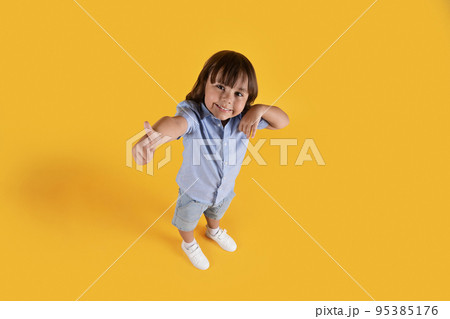 Above view portrait of little boy showing thumb up gesture, smiling to camera, orange studio background, full length 95385176
