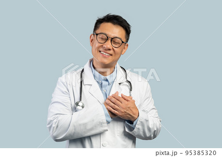 Cheerful middle aged chinese male doctor in white coat, glasses presses hands to chest 95385320