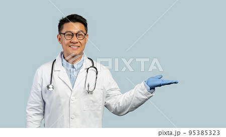 Cheerful middle aged chinese man doctor in white coat, protective gloves, goggles shows hand with empty space Cheerful middle aged chinese man doctor in white coat, protective gloves, goggles shows hand with empty space 95385323