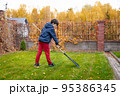 Little boy with a rake cleaning fallen leaves in the autumn yard garden. Kids and housework concept. Little boy helping with backyard cleaning. Children play outdoors. Little helper. 95386345