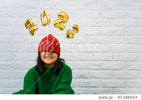 Smiling Asian baby girl in a green sweater close-up with golden balloons with numbers 2023 in hat Smiling Asian baby girl in a green sweater close-up with golden balloons with numbers 2023 in hat 95386604
