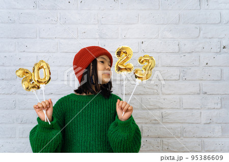 Asian cute girl in red cap and green sweater holding foil balloons with numbers 2023. 95386609