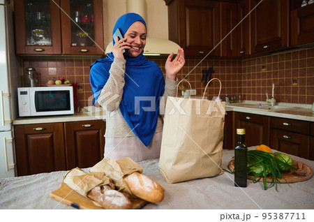 Attractive young Middle-Eastern Muslim woman with head covered in blue hijab, talking on mobile phone while arriving home after grocery shopping. Healthy food and groceries on the kitchen table 95387711