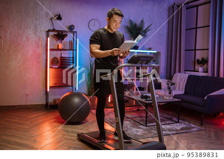 Attractive young asian man reading book working out, running, doing cardio training on treadmill 95389831