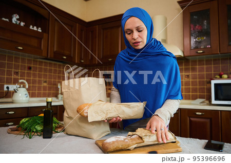 Muslim Arab woman in blue hijab, pretty housewife putting baguettes of whole grain bread on a wooden board while unpacking fresh produce from paper grocery shopping bag in the kitchen at home. 95396696