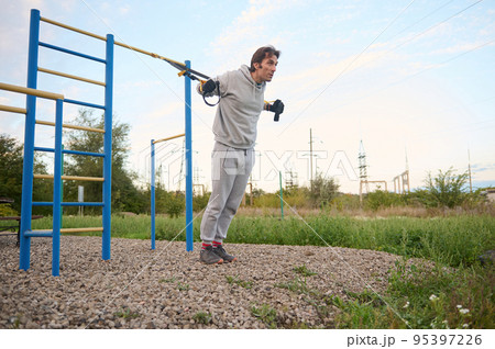 Attractive middle-aged determined Caucasian man during workout with suspension straps on the street. Athlete performs a bodyweight training on cool autumn day. Sport. Fitness. Active healthy lifestyle 95397226