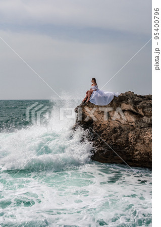A woman in a storm sits on a stone in the sea. Dressed in a white long dress, waves crash against the rocks and white spray rises above her. 95400796