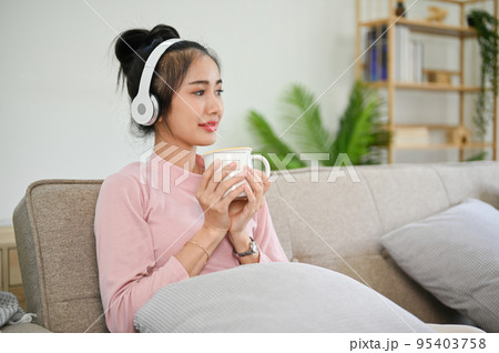 Calm Asian female listening podcast or music through her headphones while sipping tea Calm Asian female listening podcast or music through her headphones while sipping tea 95403758