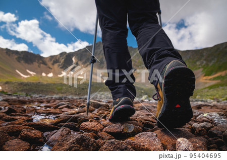Hiking boots close-up on mountain rocks by the lake with trekking poles, view from below Hiking boots close-up on mountain rocks by the lake with trekking poles, view from below 95404695