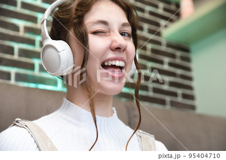 Young caucasian teenage woman smiles and winks, she listens to music in headphones while sitting at home Young caucasian teenage woman smiles and winks, she listens to music in headphones while sitting at home 95404710