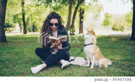 Attractive young woman is reading book sitting on grass in park while her well-bred dog is sitting near her owner and looking around. Leisure, nature and animals concept. 95407598