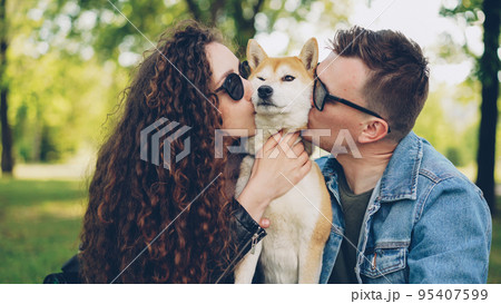 Proud dog owners pretty girl and handsome guy are playing with pet shiba inu dog, kissing it and scratching its fur relaxing in the park at weekend. People and animals concept. 95407599