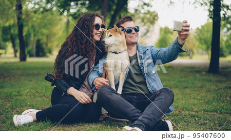 Handsome young man is taking selfie with his pretty wife and cute dog, all wearing sunglasses. Guy is holding smartphone taking funny pictures and posing. 95407600