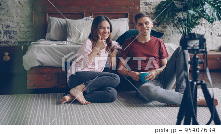 Husband and wife bloggers are recording video vlog gesturing, talking and laughing, they are using camera on tripod and sitting on bedroom floor. Husband and wife bloggers are recording video vlog gesturing, talking and laughing, they are using camera on tripod and sitting on bedroom floor. 95407634