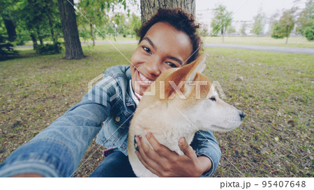 Point of view shot of pretty African American girl taking selfie with cute puppy in city park holding camera, smiling and posing. Modern technology, nature and animals concept. Point of view shot of pretty African American girl taking selfie with cute puppy in city park holding camera, smiling and posing. Modern technology, nature and animals concept. 95407648