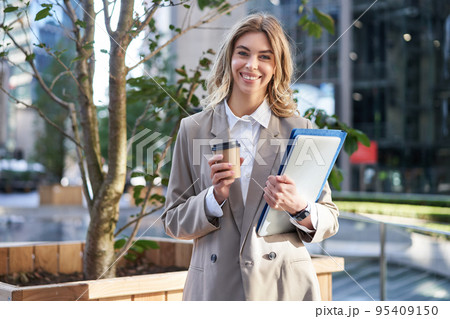 Young businesswoman standing on street with takeaway coffee, laptop and work documents, smiling Young businesswoman standing on street with takeaway coffee, laptop and work documents, smiling 95409150