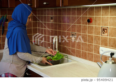 A Middle-Eastern Muslim woman, housewife in hijab, washing fresh organic vegetables by hands in the sink under running water in the home kitchen A Middle-Eastern Muslim woman, housewife in hijab, washing fresh organic vegetables by hands in the sink under running water in the home kitchen 95409421