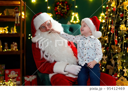 Funny boy checking the red nose of Santa Claus while sitting on his lap. New Year concept Funny boy checking the red nose of Santa Claus while sitting on his lap. New Year concept 95412469