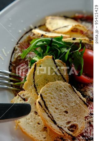 Beef Carpaccio cold appetizer with parmesan, tomatos and arugula on white plate with hardtack on dark background Beef Carpaccio cold appetizer with parmesan, tomatos and arugula on white plate with hardtack on dark background 95415429