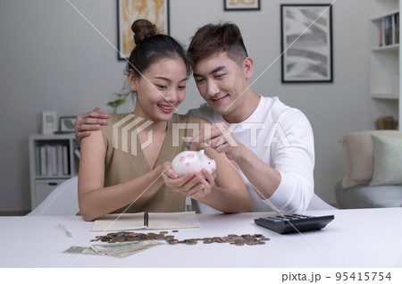 Asian couple at home saving money in a piggybank and smiling. Asian couple at home saving money in a piggybank and smiling. 95415754