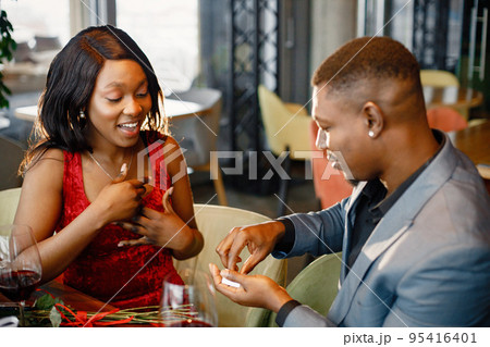 Couple enjoying day out at restaurant. Black man holding a little box with engagement ring. Woman wearing red elegant dress and man blue costume. 95416401