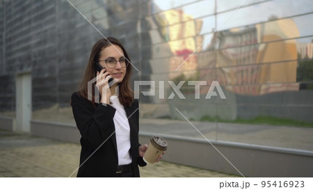 A young businesswoman is talking on the phone and drinking coffee walking around the business center. A woman in glasses and a suit is on her lunch break. A young businesswoman is talking on the phone and drinking coffee walking around the business center. A woman in glasses and a suit is on her lunch break. 95416923