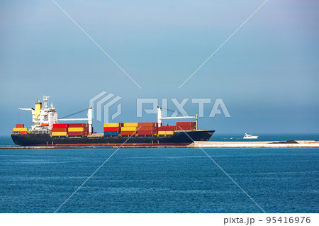a bulk carrier with cranes for unloading cargo transports multi-colored containers, a small-sized sea vessel sails along the sea along the breakwater next to the port against the sky on a sunny day. a bulk carrier with cranes for unloading cargo transports multi-colored containers, a small-sized sea vessel sails along the sea along the breakwater next to the port against the sky on a sunny day. 95416976
