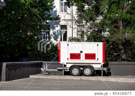 White and red Backup Generator on the trailer with wheels. Mobile emergency Generator or Standby Generator - Outdoors Power Equipment on the street nearby trees and building on sunny day, nobody. 95416977