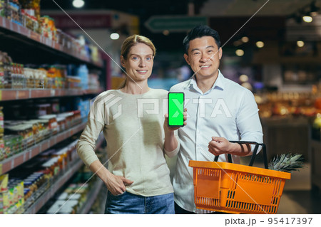 Young family diverse couple of shoppers in supermarket, smiling and looking at camera, grocery department, man and woman holding shopping basket and showing green screen of smartphone. 95417397