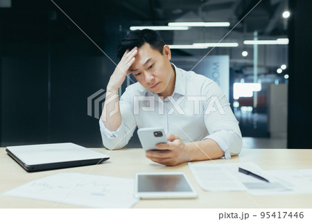 Confused Asian guy holding smart phone feels concerned thinking over received message. Mobile phone everyday usage, unpleasant news, waiting for important call, low signal device problems concept 95417446