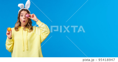 Holidays, traditions and celebration concept. Amused lovely european female blonde in rabbit ears, look excited and surprised as holding two painted Easter day eggs, blue background 95418947