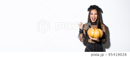 Image of excited beautiful asian woman celebrating halloween, wearing witch outfit and holding pumpkin, yelling with rejoice 95419026