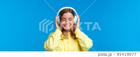 Music, lifestyle and youth concept. Close-up portrait of dreamy happy and relaxed young blond european girl close eyes and enjoying fine sound of song in headphones, dreaming, blue background 95419077