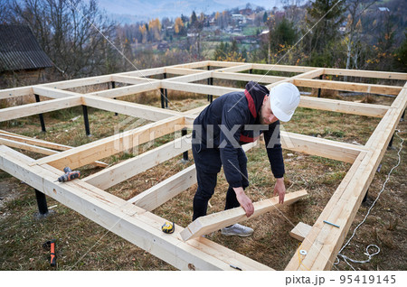 Men workers building wooden frame house on pile foundation. Carpenters installing wooden truss for timber framing. Carpentry concept. Men workers building wooden frame house on pile foundation. Carpenters installing wooden truss for timber framing. Carpentry concept. 95419145
