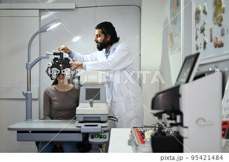 Male ophthalmologist with beard in white examining checking eye vision of patient with phoropter eyesight measurement testing machine Male ophthalmologist with beard in white examining checking eye vision of patient with phoropter eyesight measurement testing machine 95421484