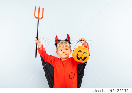 Cute boy in devil halloween costume with trident and pumpkin basket 95422556