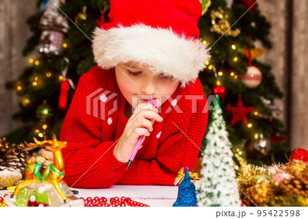 Boy in Santa hat writes letter to Santa Claus near Christmas tree 95422598